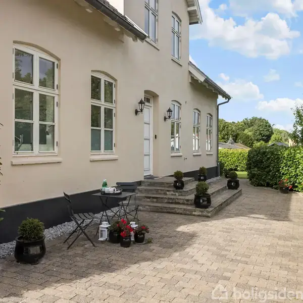 En beige bygning med hvide vinduer står ved en flisebelagt gårdhave. To sorte caféstole og et bord med drikkevarer er omgivet af potteplanter. Grønne hække ses i baggrunden.