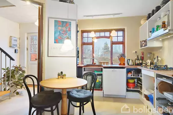 Rundt bord med to stole står i et lyst køkken med vindue, hylder og farverige køkkenudstyr. En plante og trappe ses i baggrunden. Et billede hænger på væggen.