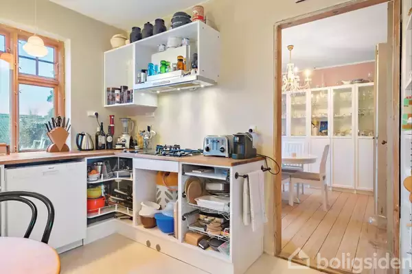 Et veludstyret køkken viser åbne skabe med køkkenudstyr og en bordplade med forskellige apparater, som står ved siden af en lys spisestue med store skabe i baggrunden.