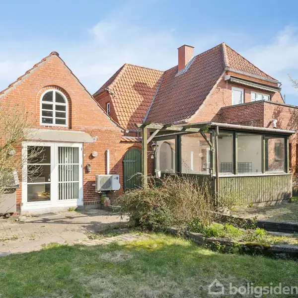 Rødt murstenshus med buede vinduer og glasudestue. Huset har en velplejet have med græsplæne og buske. Solrigt vejr giver en lys, indbydende atmosfære.