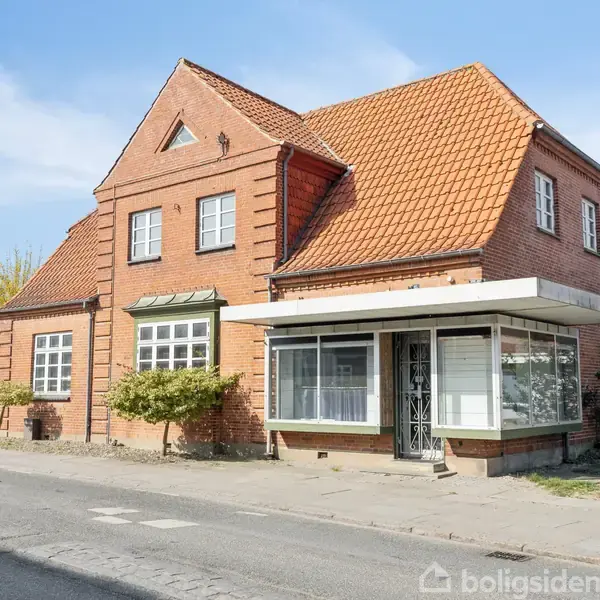 En murstensbygning med rødt tegltag står ved en vej. Den har store vinduer, nogle med markiser, og en indgangssektion med glas. Omgivet af buske og træer.