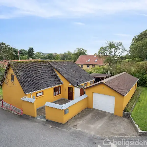 Et gult hus med mørkt tag står ved vejen. Ved siden af er en garage og en grøn græsplæne. Omkring er der grønne træer og en anden bygning ses i baggrunden.