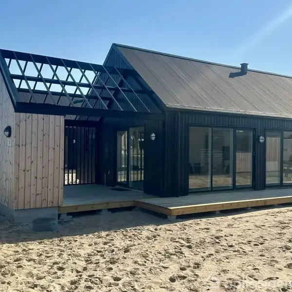 Et moderne feriehus med skråt tag og store glaspartier står på sandet grund. Teksten på billedet siger: "Modelfoto fra tilsvarende feriehus."