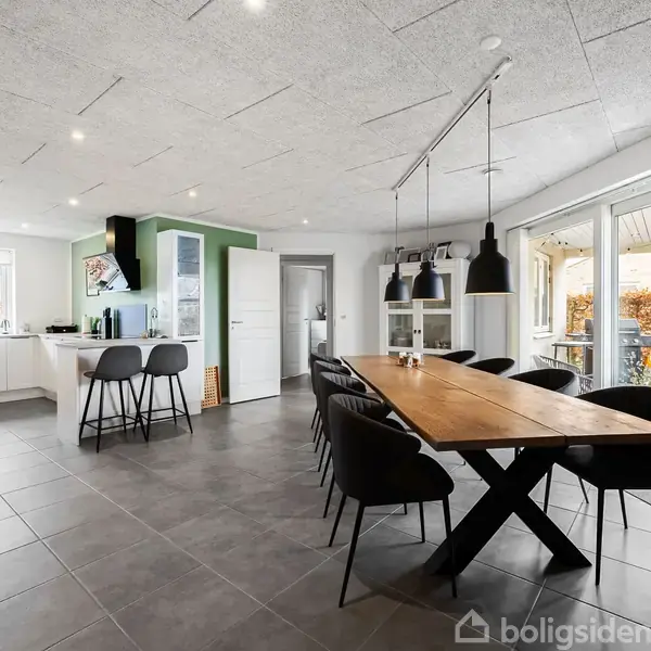 Et rummeligt, moderne køkken-alrum med et stort træbord omgivet af sorte stole. Køkkenet inkluderer en ø med barstole. Store vinduer og døre åbner til verandaen udenfor.