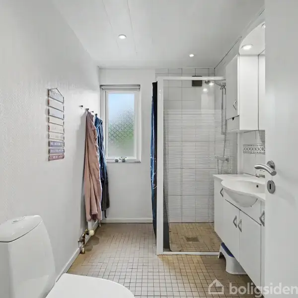 Et badeværelse med et toilet i forgrunden. Brusekabine og vaskenhed er på højre side. Væggene er lyse, og der hænger håndklæder ved et vindue i baggrunden.