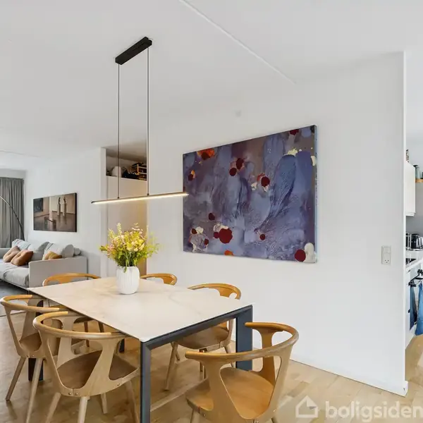 En lys spisestue har et bord med seks træstole og en vase med blomster på. På væggen hænger et abstrakt maleri. I baggrunden ses en stue med sofa.