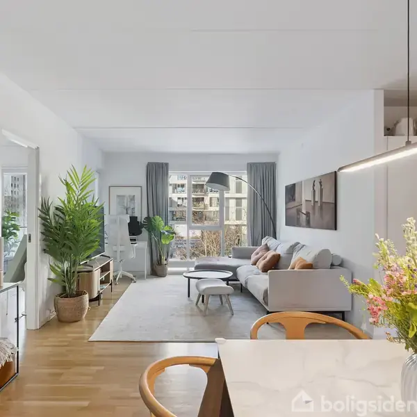 Stue med en grå sofa og et rundt sofabord, placeret på et tæppe. Store vinduer bagved slipper lys ind. En spisestol og bord ses foran blomster i vasen.