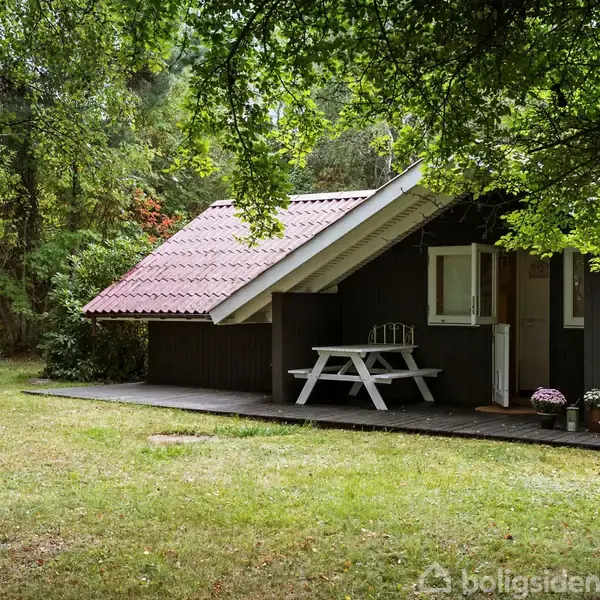 Hyggeligt sommerhus med rødt tegltag står i en frodig skov. En hvid bænk står foran huset på en træterrasse, omgivet af grønne græsplæner og tætte træer.