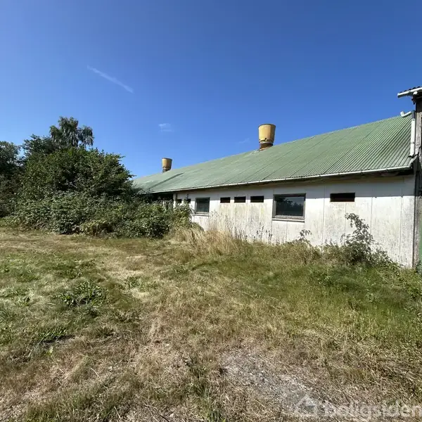 Et hvidt, to-etagers hus står midt i en græsplæne med solskin og få træer omkring. Til venstre ses en ældre bygning. Klar blå himmel over det hele.