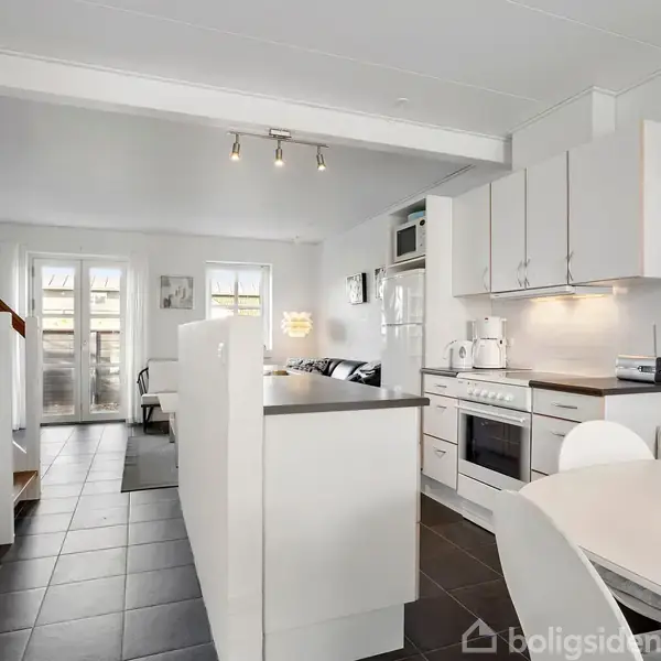 Køkken-alrum i moderne stil med hvide elementer. Køkkenøen og spisebordet er centralt placeret. I baggrunden ses stue med lyse vægge og store vinduer, der lukker lys ind.