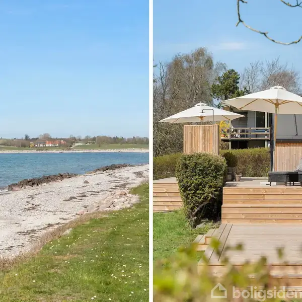 Strand med sand og græs langs kysten. Terrasse med liggestole og parasoller foran træhus. Blandet miljø af natur og bolig ved vandet under en klar blå himmel.