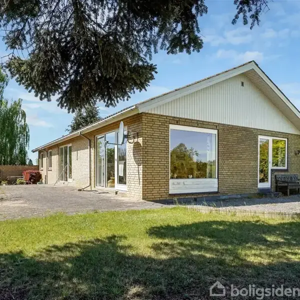 En étplans villa med store vinduespartier står i en grøn have med en flisebelagt terrasse. Der er træer og buske i baggrunden under en blå himmel med spredte skyer.