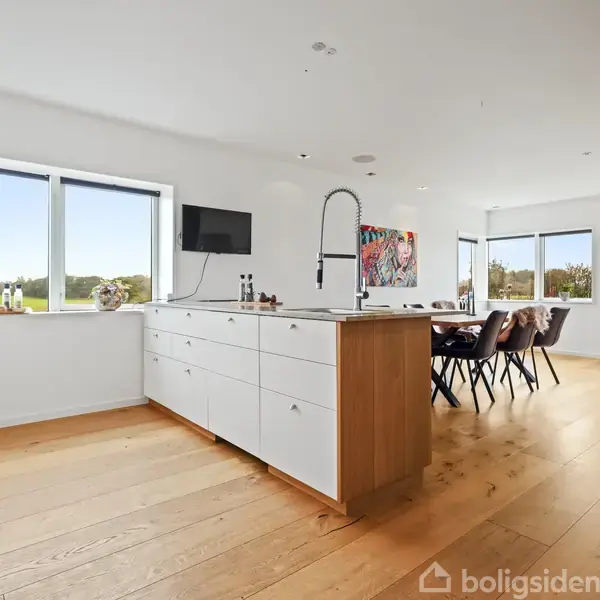 Et moderne køkken har et stort hvidt køkkenbord med integreret vask. I baggrunden står et spisebord med stole. Rummet har trægulv og store vinduer med udsigt til grønt landskab.