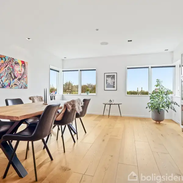 Et træbord med sorte stole står i et lyst rum med trægulv. Væggene har kunst og store vinduer, og der er en plante ved siden af et vitrineskab.