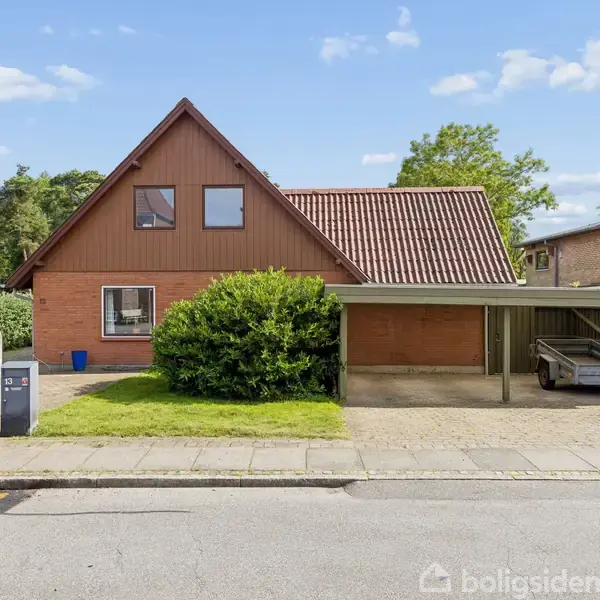 Brunt etplanshus med skråtag, placeret i en forstad. Huset har en carport med en trailer. En grøn busk vokser foran huset, og der er blå himmel.