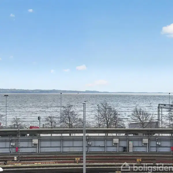 Udsigt over en jernbanestation med skinner i forgrunden, træer og en havudsigt i baggrunden under en klar blå himmel med spredte skyer.