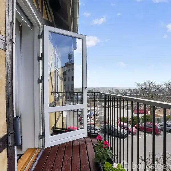 Balkon med åben havedør, udsigt over parkeringsplads og bygninger i baggrunden. Rækværket har blomsterdekorationer, og vejret ser klart ud med blå himmel og få skyer.