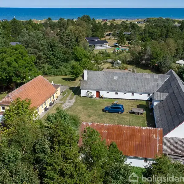 Store gårdbygninger omkranser en gårdsplads med en blå bil parkeret. Bygningerne er placeret i et grønt, skovrigt område tæt på havet i baggrunden.