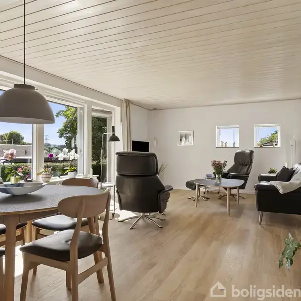 Moderne stue med spisebord og stole foran store vinduer, der åbner for udsigt til en terrasse med blomster. Behagelige lænestole og sofa står i en lys og rummelig indretning.