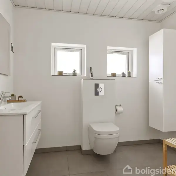 Toilet hænger på væggen; omgivet af lys indretning med to små vinduer over det. Til venstre står en hvid håndvask med spejl, flasker og skab.