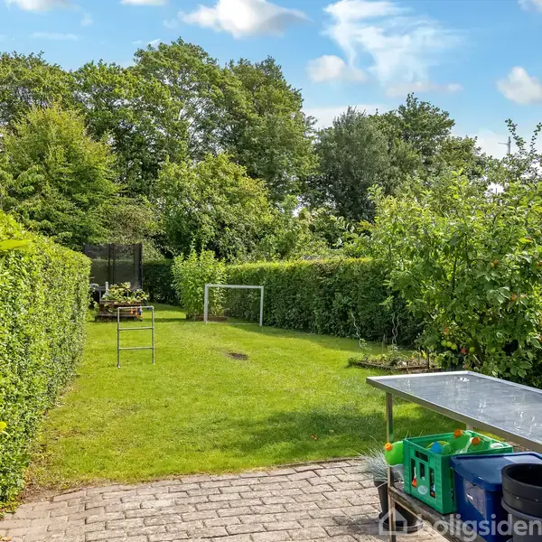 Græsplæne omgivet af hække med et fodboldmål og bordtennisbord i baggrunden, placeret i en frodig have med blå himmel og spredte skyer.