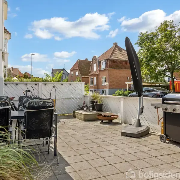 Terrasse med bord og stole, parasol og grill. Omgivet af et træhegn, med udsigt til røde murstenshuse og træer på en solrig dag.