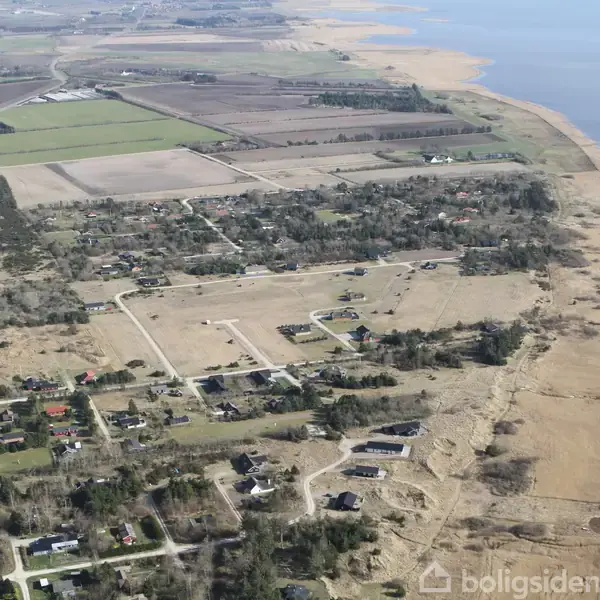 Luftfoto viser et kystnært område med spredte huse og veje omgivet af marker. Kystlinjen strækker sig langs højre side, og landskabet er fladt og grønt.