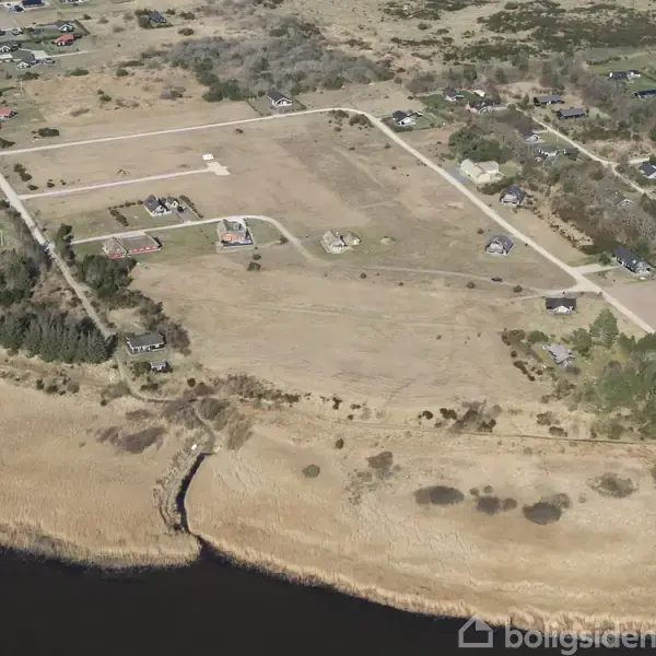 Et fugleperspektiv viser et landligt område med spredte huse og veje omgivet af åbne marker. En kystlinje og vand kan ses i forgrunden.