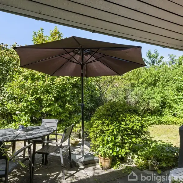 En parasol står over et rundt havebord med stole. Sollyset skinner på træterrassen omkranset af frodige buske og grønne planter i en hyggelig baghave.