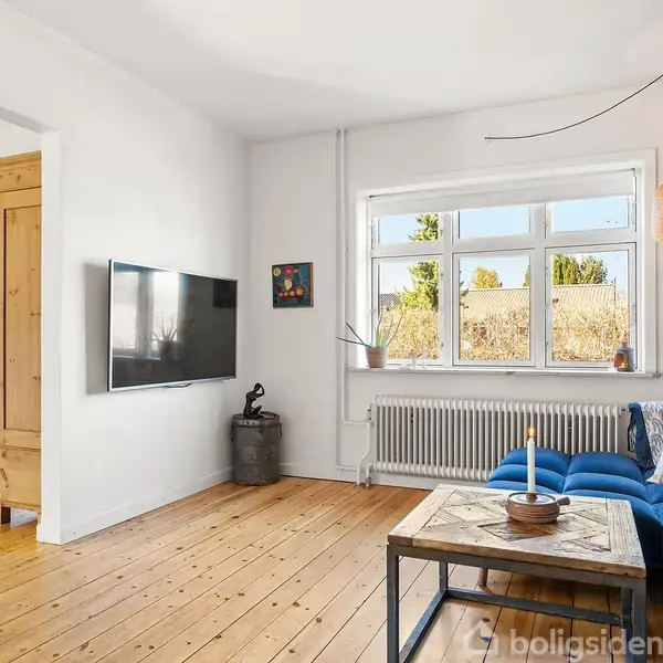 Stue med blå sofa og sofabord, fladskærms-tv på væggen. Vindue med udsigt til have, orange lampe hænger fra loftet. Til venstre en åbning til spiseområde.