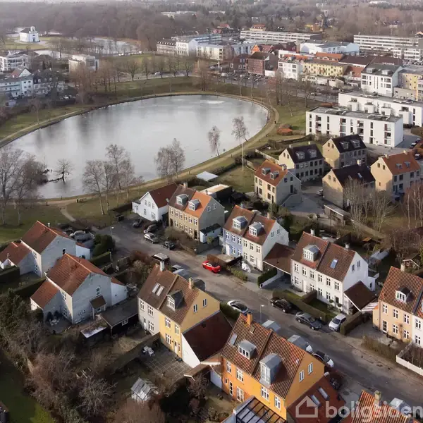 En række huse ligger langs en vej i et boligområde. Bagved ses en rund sø omgivet af træer og flere bygninger i baggrunden.