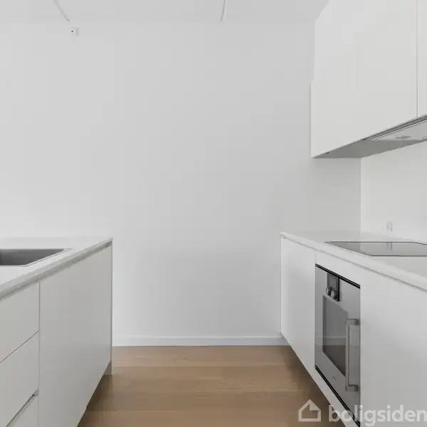 Et moderne, minimalistisk køkken med hvide skabe og en integreret ovn. En stålvask er placeret i en fritstående bordplade til venstre. Trægulve kompletterer det lyse rum.