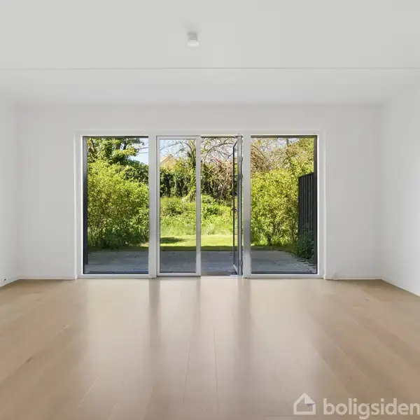 Et rummeligt, lyst værelse med trægulv og en stor glasdør der åbner ud mod en frodig have. Væggene er hvide, og der er et minimalistisk udtryk.