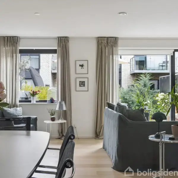Stue med en grå sofa står ved siden af store vinduer, som giver udsyn til en terrasse fyldt med planter. Et spisebord med blomster synes i forgrunden.