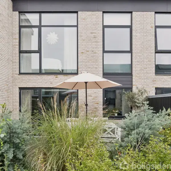 En beige parasol står i en have foran et moderne murstenshus med store vinduer. Omkring parasollen er der grønne planter og buske.