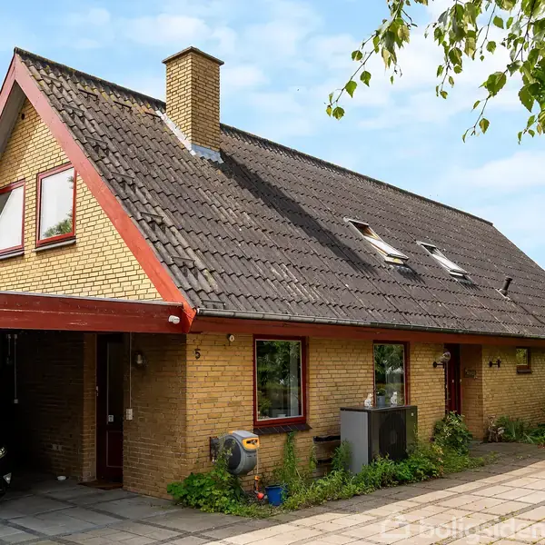 Et gult murstenshus med mørkt skråtag står ved siden af en carport, hvor en bil er parkeret. Nummeret "5" er synligt ved hoveddøren. Grønne planter omgiver huset.