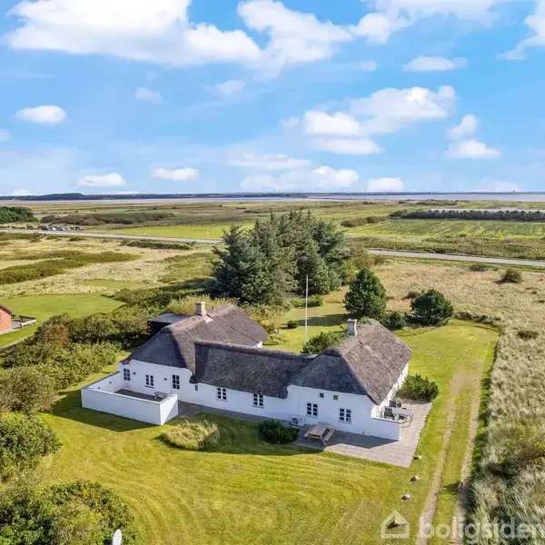Hvidt hus med stråtag står stille midt i et grønt landskab med klitter og få træer i nærheden, under en åben blå himmel med skyer.