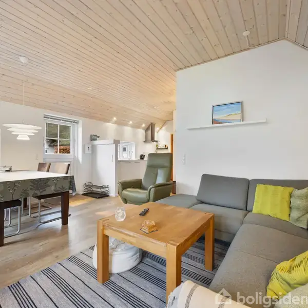 Grå hjørnesofa står i en lys stue med et træbord foran, i et rum med trægulv og -loft. Spisebord med stole ses ved stort vindue.