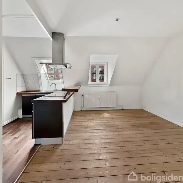 Et moderne køkken med sorte skabe og en indbygget ovn. Overbordet hænger en emhætte. Rummet har hvidmalede vægge, et skråvindue og trægulv. En radiator er under vinduet.