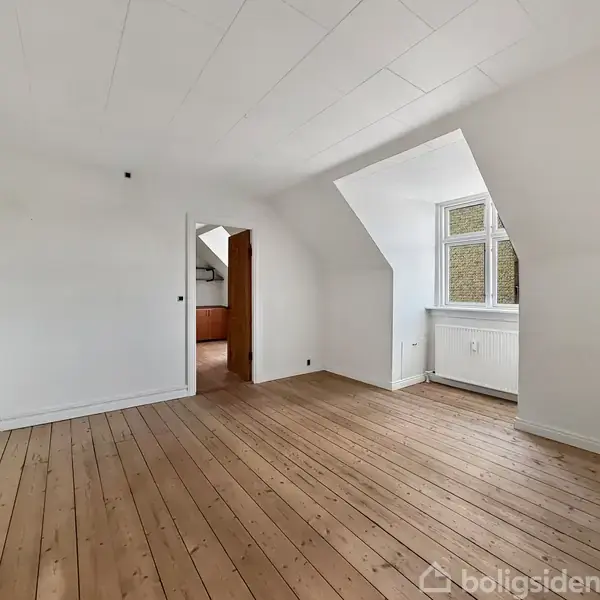 Et tomt rum med bræddegulv, en hvidmalet væg og et skråloft. Der er en dør til et andet rum, og et vindue med udsigt til en murstensvæg.