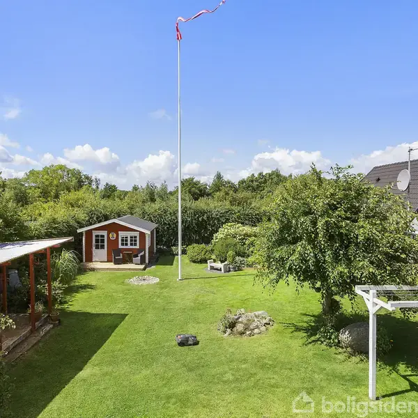 En have med et græsareal omgivet af buske og træer. En flagstang står midt i haven, mens et lille træhus ses bagerst. En hvid pergola står til højre.