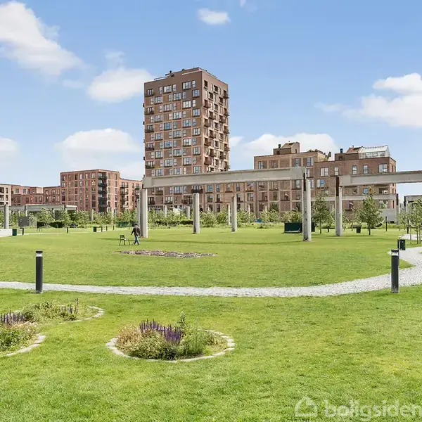 Grøn parkareal med stier og blomsterbede omkranset af moderne lejlighedsbygninger. En høj søjlegang forbinder områderne, mens blå himmel og skyer ses i baggrunden.