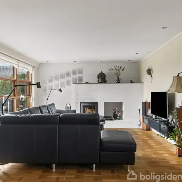 En brun murstensvilla med sort tag står i en velholdt have. Der er en ældre vandpumpe på en lille stenbed foran huset, omgivet af grønne buske og træer.