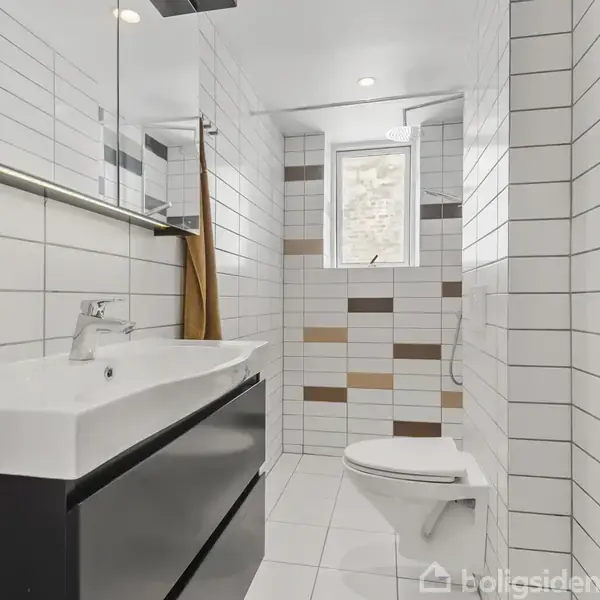 Håndvask med moderne spejl hænger på væggen i et lyst badeværelse med hvide og brune fliser; et toilet står nær et vindue.