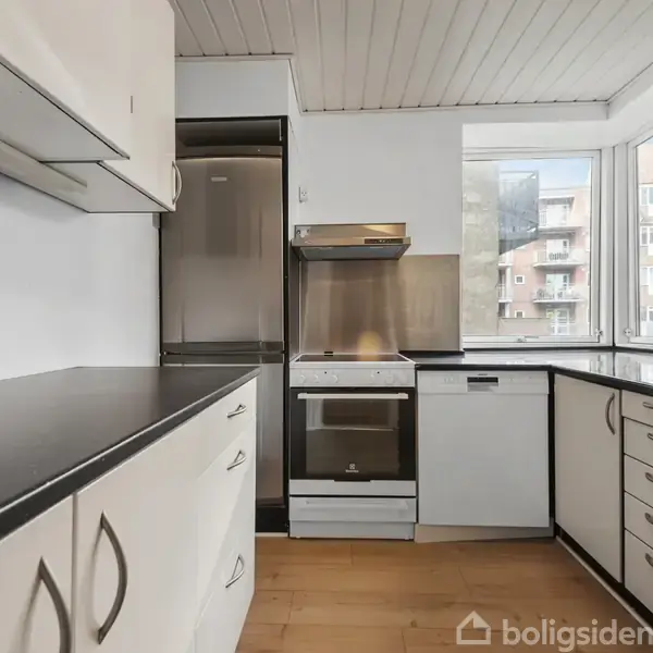 Et moderne køkken med hvide skabe og sorte bordplader dominerer rummet. En ovn med emhætte ses midtfor, flankeret af et køleskab. Store vinduer giver udsigt til bygninger udenfor.