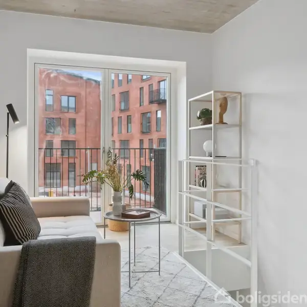 Sofa står foran store vinduer med altanudsigt mod røde murstensbygninger. En hylde med dekorationer til højre, og en gulvlampe samt sofabord i midten fuldender indretningen.