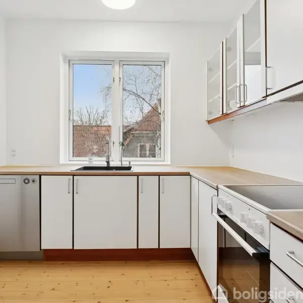 Køkken med hvide skabe og træbordplader. En opvaskemaskine og en komfur står langs væggene. Et vindue giver udsigt til træer og hustage.