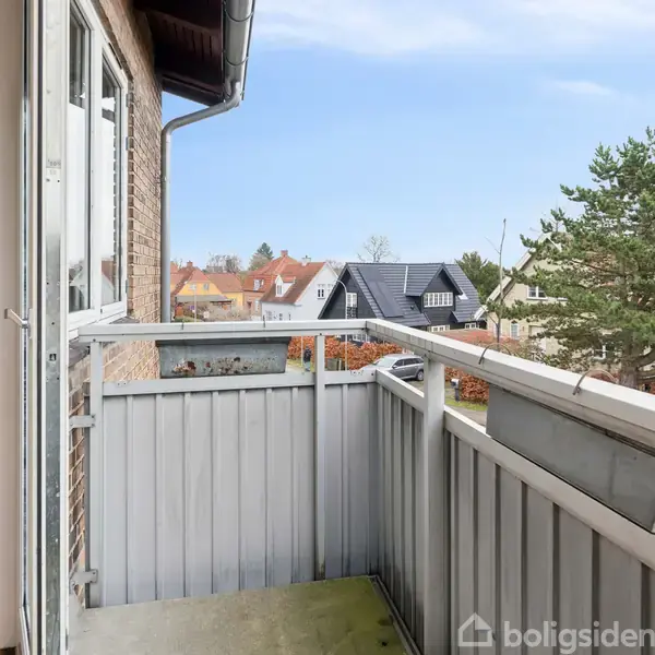 En lille balkon med hvidt rækværk vender ud mod et boligområde med hustage og grønne træer. Sidedør fra et murstenshus åbner ud til balkonen.