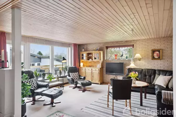 Stue med lænestole og sofa arrangeret rundt om et sofabord, foran en tv-skærm. Store vinduer giver udsigt til naboens hus. Rummelige hylder og planter pynter rummet.