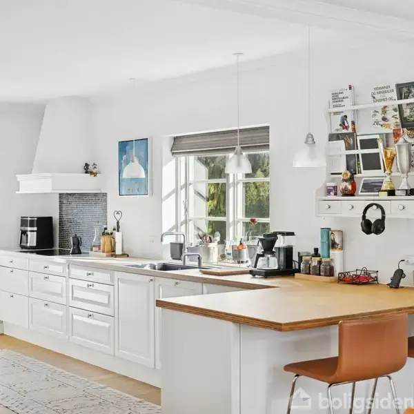 Et lyst køkken med hvid indretning. Bordpladen er dekoreret med kaffemaskiner og krydderibeholdere. To brune stole står ved køkkenøen. Et opslagstavle hænger på væggen.